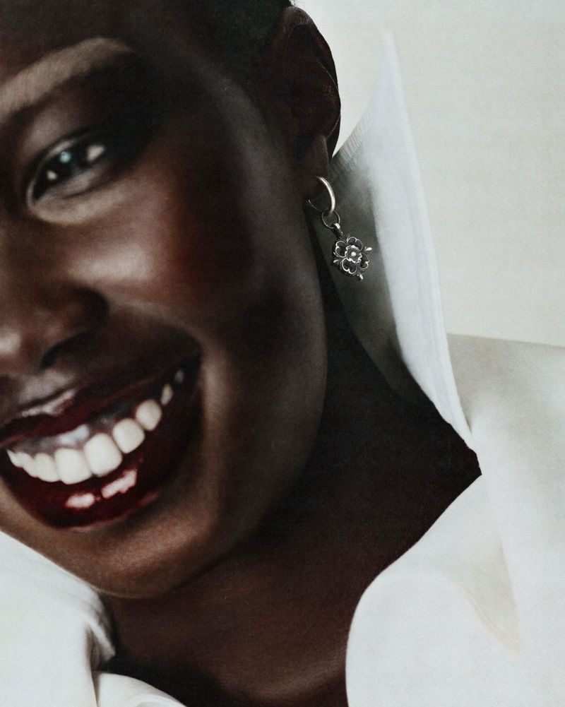 Close-up portrait of a smiling Black woman wearing a crisp white shirt and small silver charm earring against a neutral backdrop.