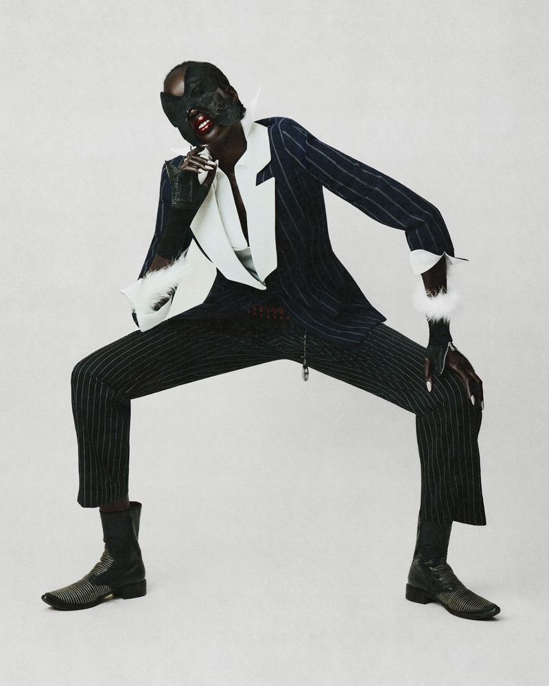 Model in a black pinstripe suit with white lapels and cuffs wearing a dark mask and boots against a light grey background.