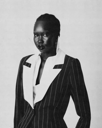 Black woman wearing a structured pinstripe suit with a large white lapel collar against a plain neutral background.
