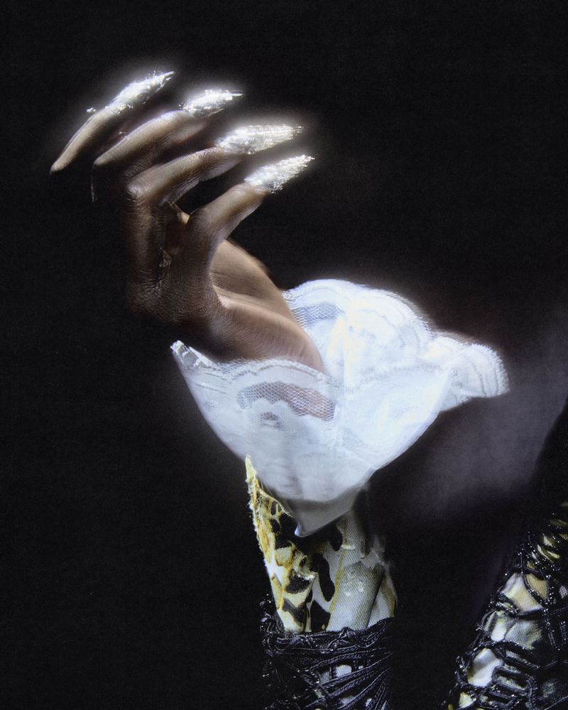 Close-up of a person with dark skin holding up a hand with long glittery fingernails and wearing a white lace ruffled cuff.
