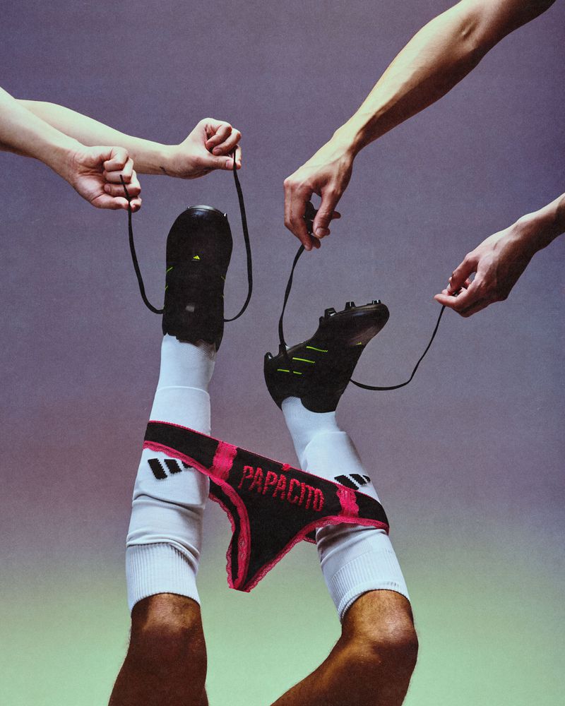 Close-up of legs lifted up wearing soccer cleats, white socks, and a black thong hung over the shins, with hands holding the laces against a grey studio backdrop.
