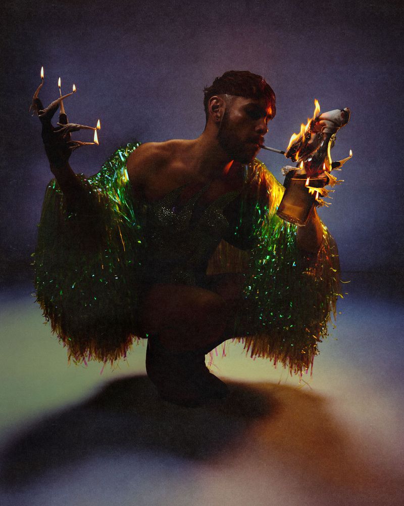 Man crouching in a shimmering green fringe jacket, with lit candles on his fingers and holding a burning object in a dark studio.