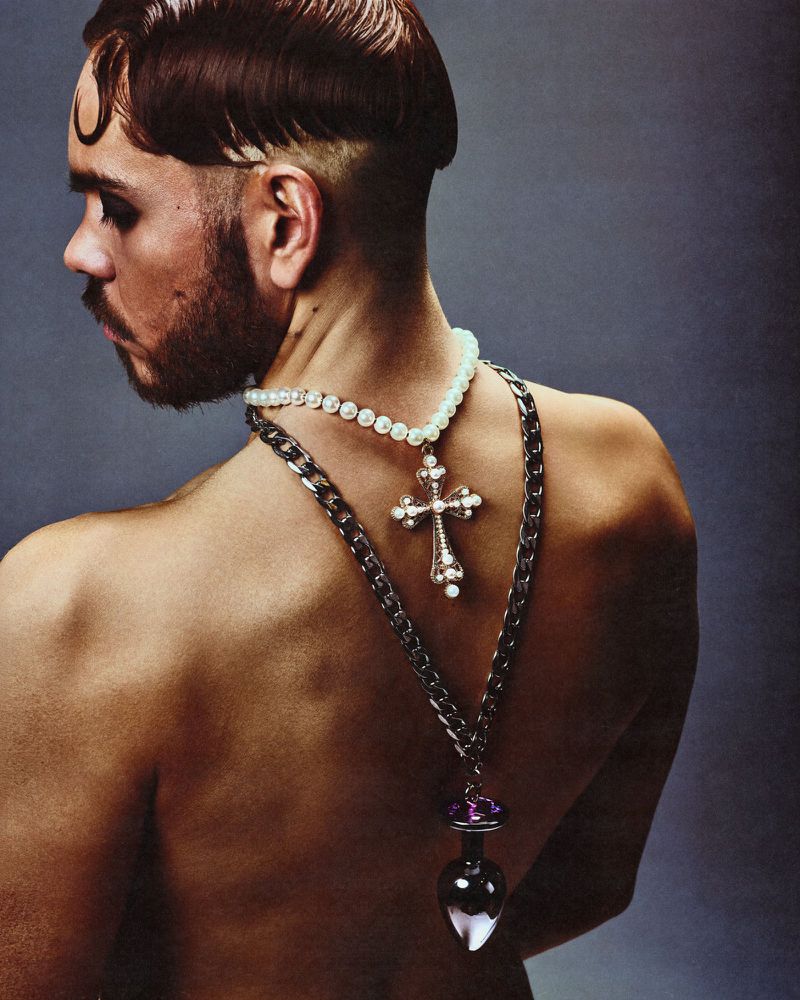 Profile portrait of a shirtless man wearing layered cross and gemstone necklaces against a dark grey background.