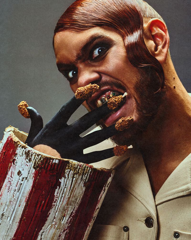 Close-up portrait of a man with styled reddish-brown hair biting into fried chicken pieces placed on his black-painted fingers for Fault Magazine
