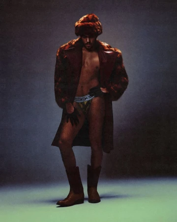 Man wearing a fur hat, fur coat, briefs, gloves, and cowboy boots against a textured grey background.