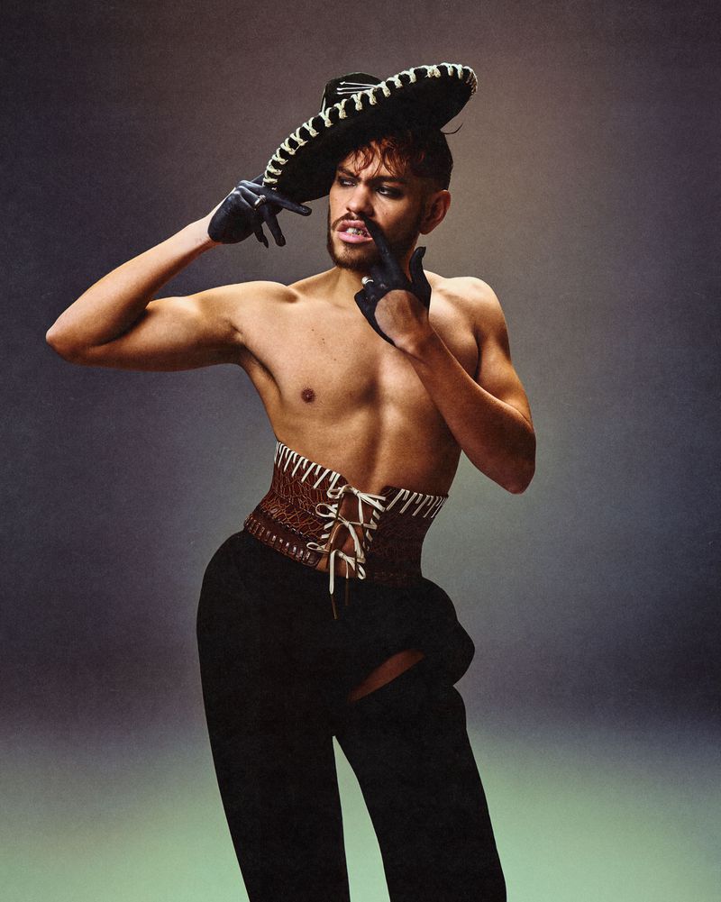 Shirtless man wearing a decorated wide-brimmed hat, laced corset, and dark trousers against a neutral studio backdrop