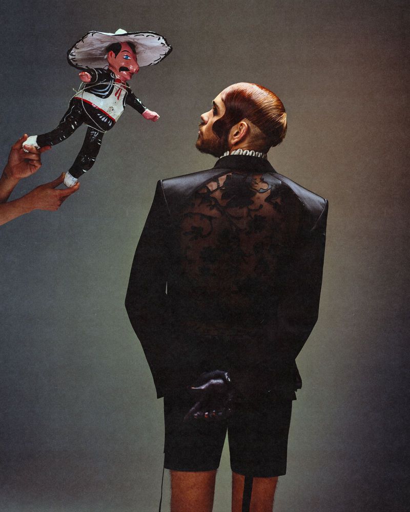Adult male in black suit looks at marionette in a sombrero held by unseen hands against a muted grey background in studio lighting.
