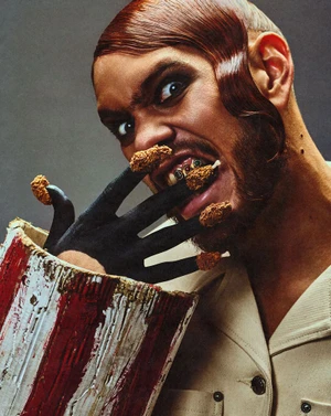 Close-up portrait of a man with styled reddish-brown hair biting into fried chicken pieces placed on his black-painted fingers for Fault Magazine