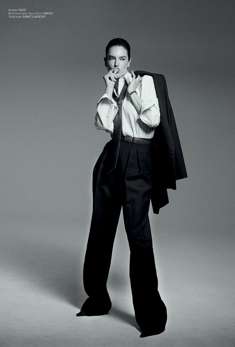 Alessandra Ambrosio posing in a white button-up shirt, black tie, and high-waisted wide-leg trousers with a blazer slung over her shoulder.