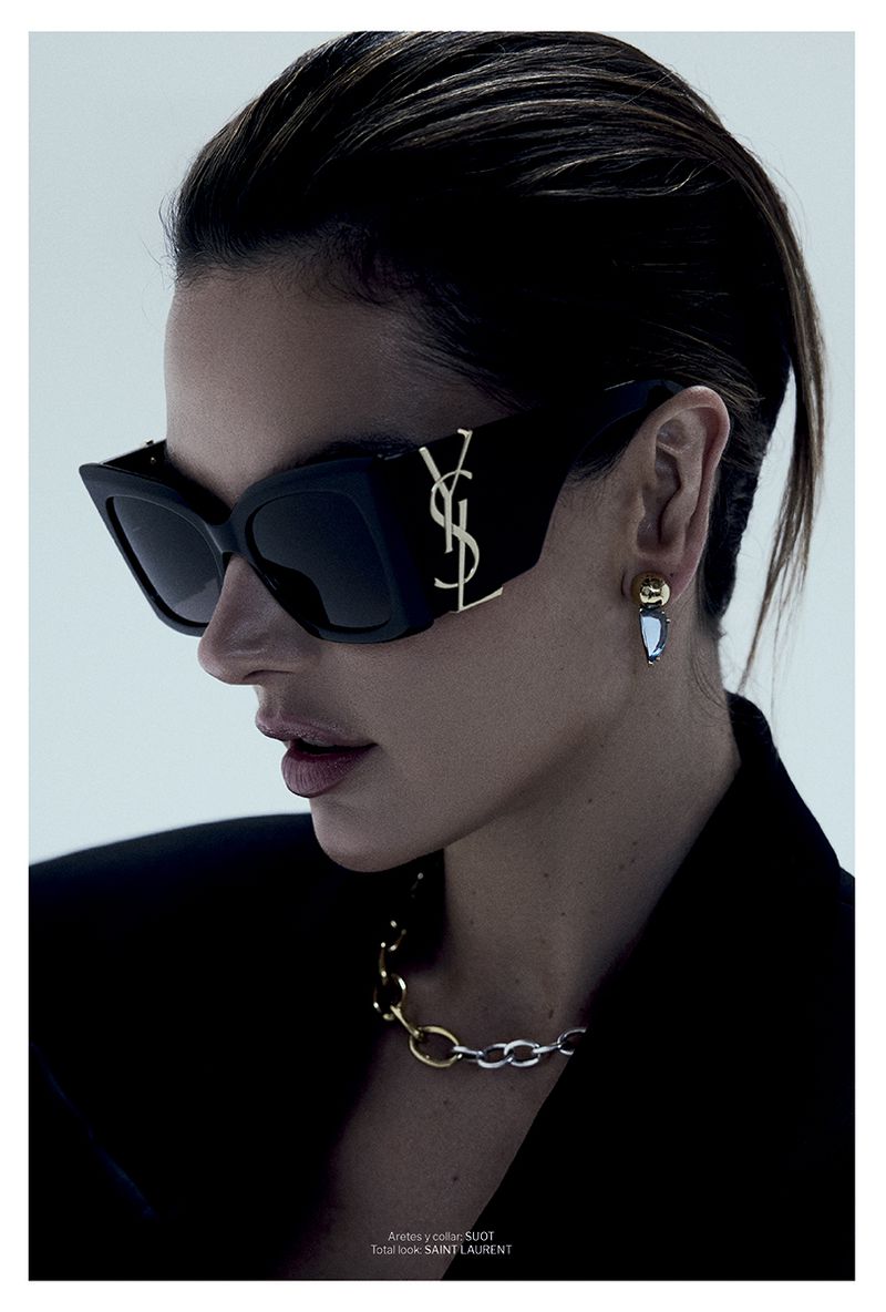 Alessandra Ambrosio wearing black Saint Laurent sunglasses with a gold logo, chain necklace, and black blazer against a grey backdrop.