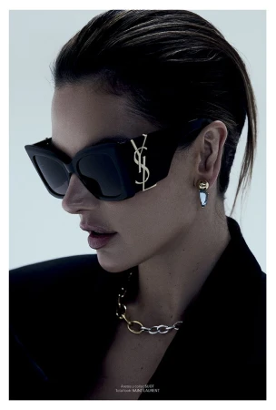 Alessandra Ambrosio wearing black Saint Laurent sunglasses with a gold logo, chain necklace, and black blazer against a grey backdrop.