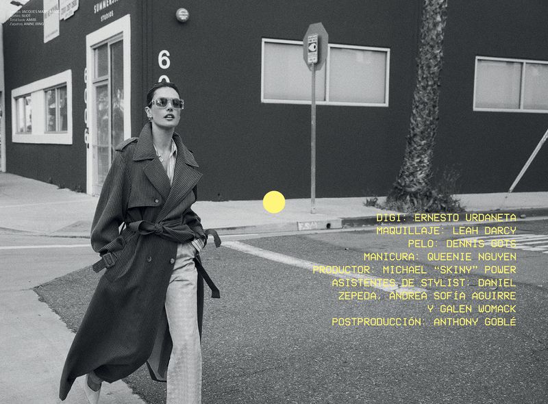An adult woman is walking on a city street. She is wearing a trench coat, sunglasses, and trousers. A building is in the background.