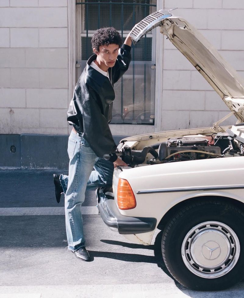 Model in a black leather jacket and blue jeans posing with a vintage car with its hood open on a street.