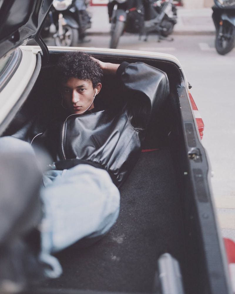 Young man wearing a black leather jacket lying inside the open trunk of a car on a street