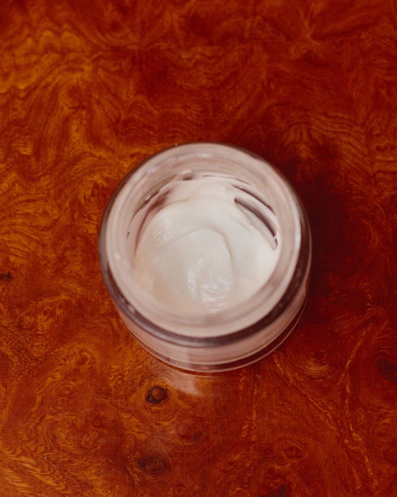 Top-down view of an open glass jar filled with white skincare cream resting on a rich brown wood grain surface.