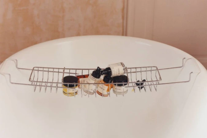 Several skincare bottles arranged in a metal caddy resting across a white porcelain bathtub.
