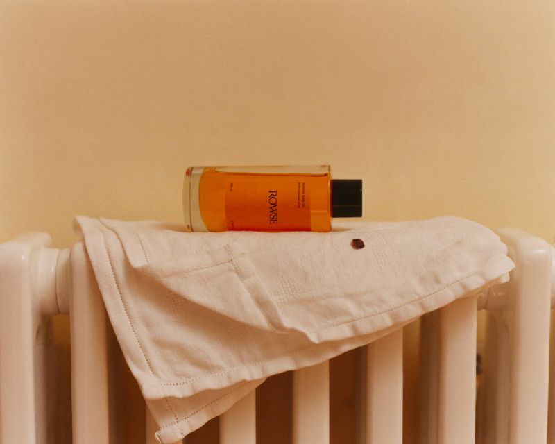 An orange glass bottle of body oil resting on a white folded towel over a radiator