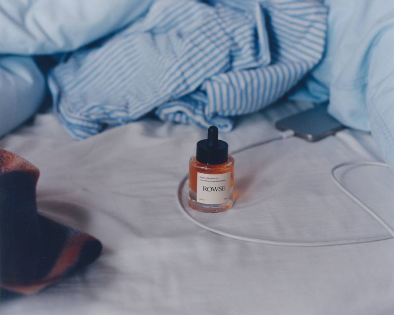 Rowse skincare dropper bottle resting on rumpled white bedsheets with blue striped pyjamas