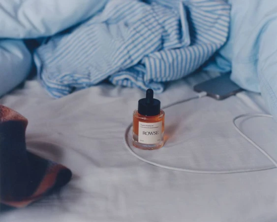 Rowse skincare dropper bottle resting on rumpled white bedsheets with blue striped pyjamas