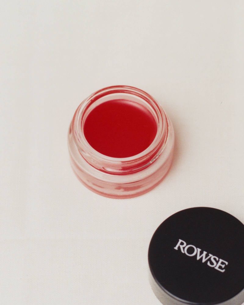Open glass cosmetics jar filled with red lip tint next to a black cap with white Rowse branding on a neutral surface