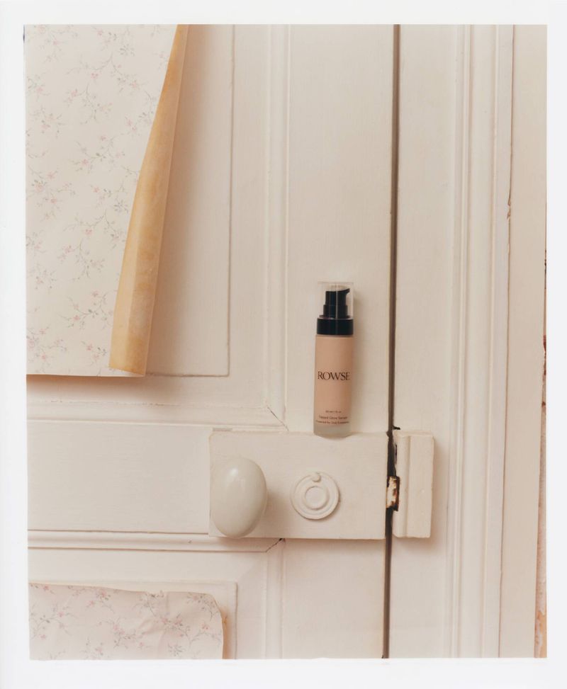 A bottle of Rowse face serum set against a white wooden door, complemented by floral wallpaper with a beige geometric paper accent.