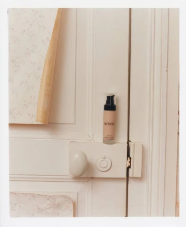 A bottle of Rowse face serum set against a white wooden door, complemented by floral wallpaper with a beige geometric paper accent.