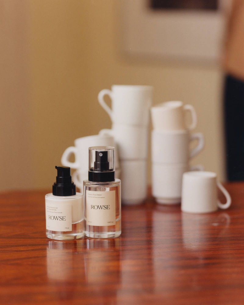 Two glass cosmetic bottles with black pumps sit on a polished wooden table with stacked white mugs in the background.