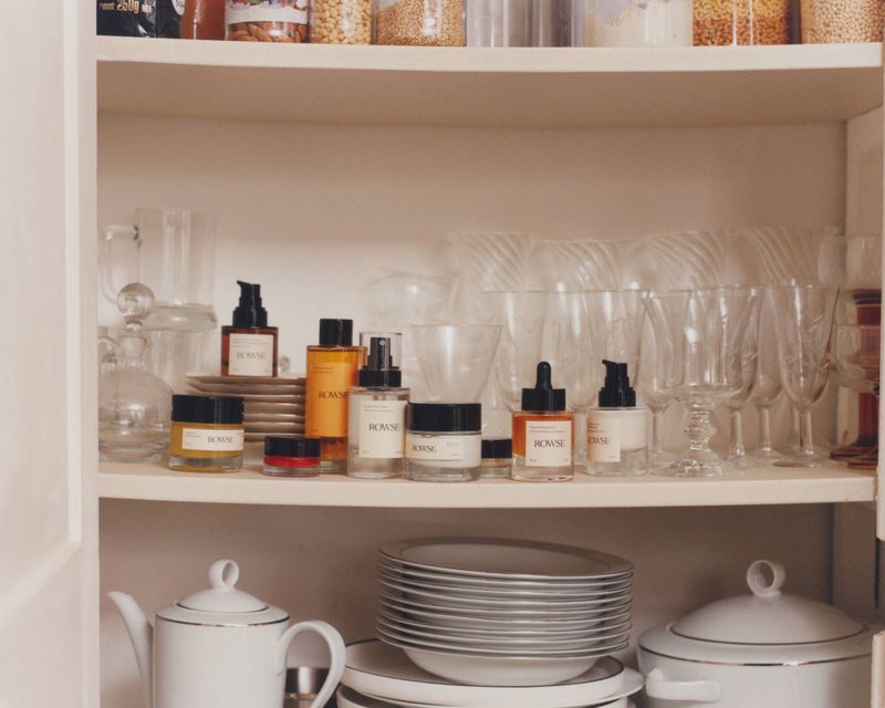 Rowse skincare and beauty products arranged neatly on shelves inside a white cupboard alongside glassware and porcelain dishes.