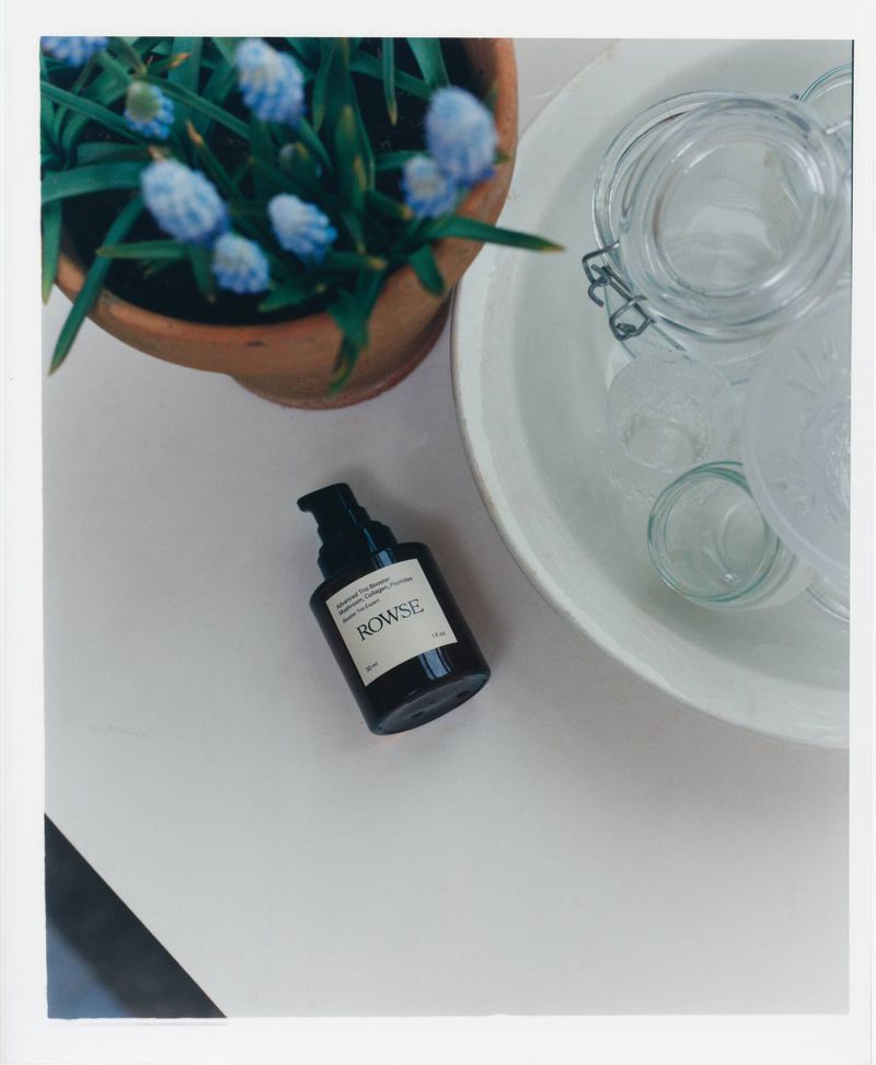 Rowse skincare product bottle next to a potted plant and glass jars on a white surface