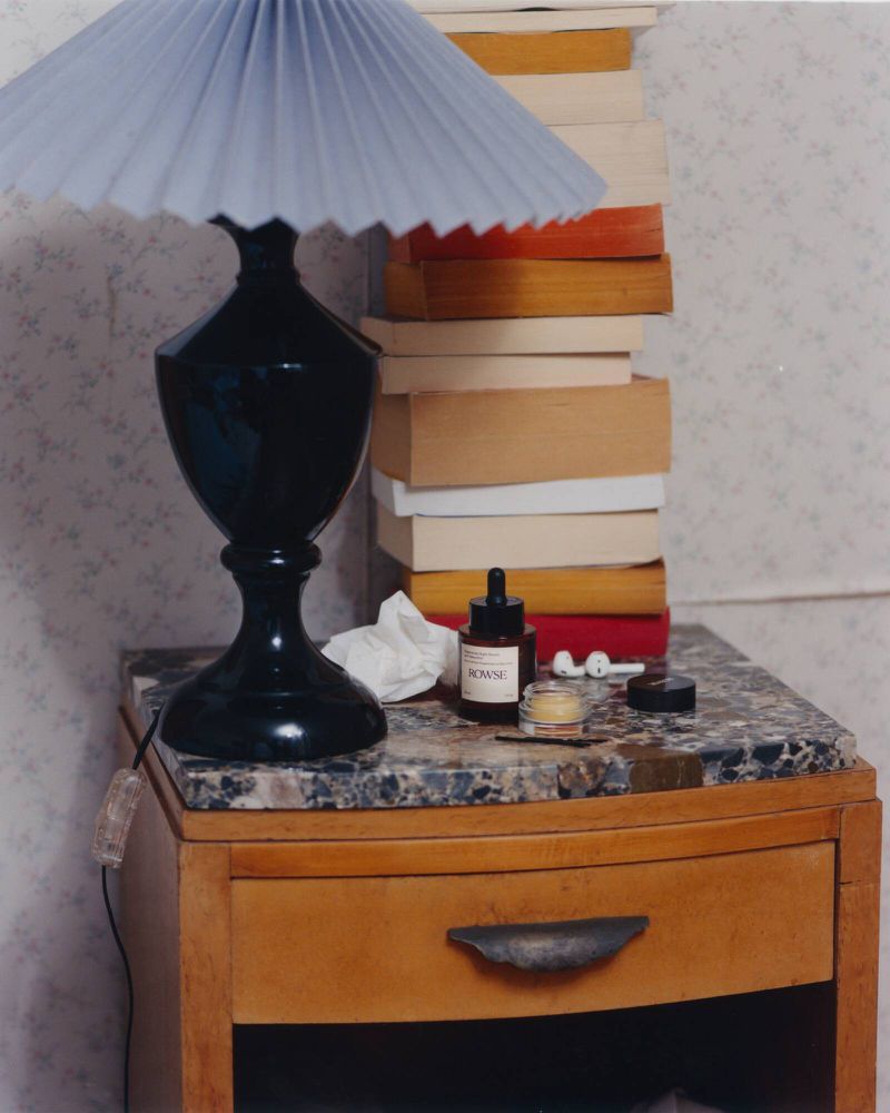Black pleated lamp and Rowse skincare products on a marble-topped wooden nightstand with stacked books.