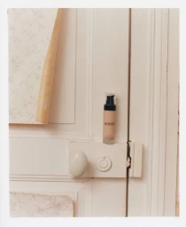 Rowse skincare pump bottle resting on a white painted door latch next to peeling floral wallpaper.