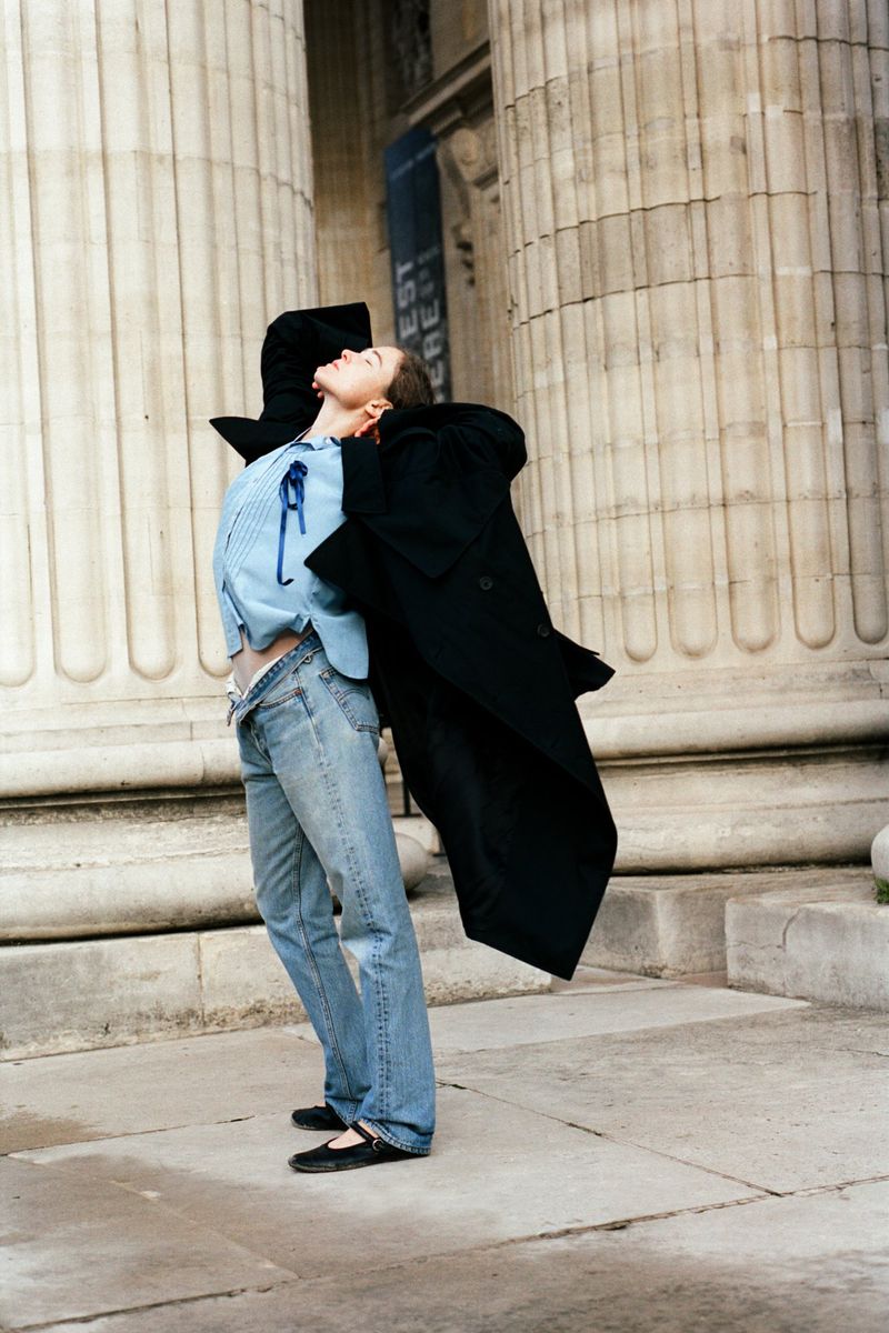 Model leaning back against massive classical columns in a light blue shirt, denim jeans, and a flowing black coat.