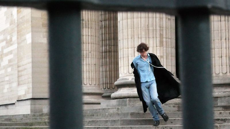 Man wearing a light blue shirt and dark coat walking down stone steps in front of classical columns