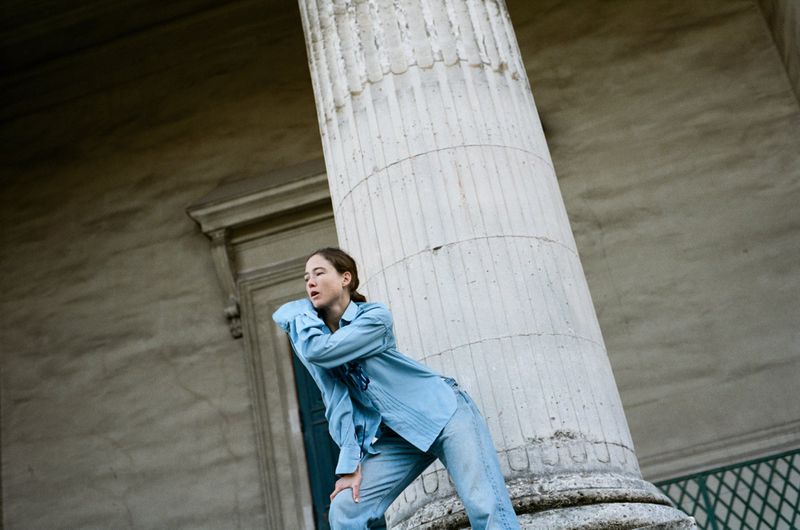 Young woman in a blue shirt and jeans poses next to a large fluted column outdoors in front of a building with a stone facade.