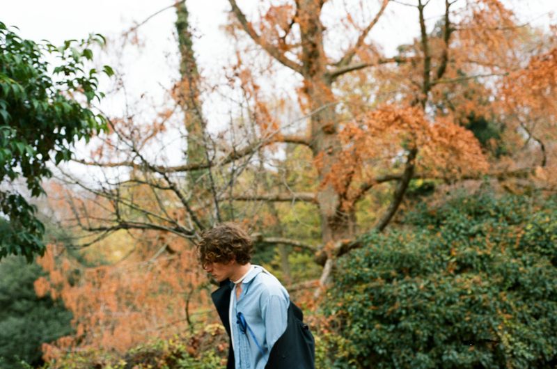 Man wearing a light blue shirt with a dark jacket draped over his shoulders standing outdoors in a wooded park.