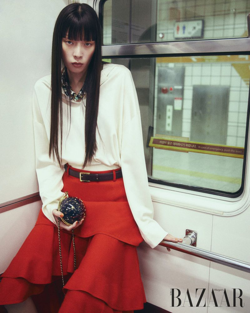 Model with long dark hair wearing a white top and red skirt seated inside a train carriage