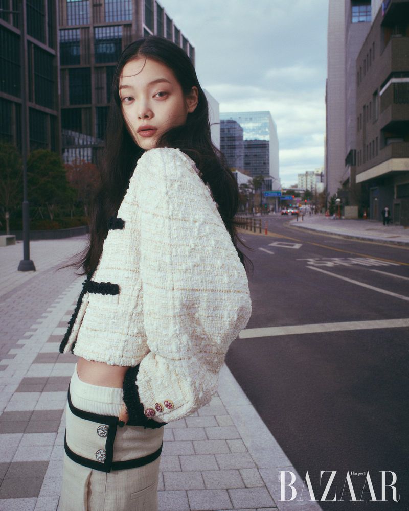 Model wearing a textured white jacket and skirt standing on a city street beside a road.