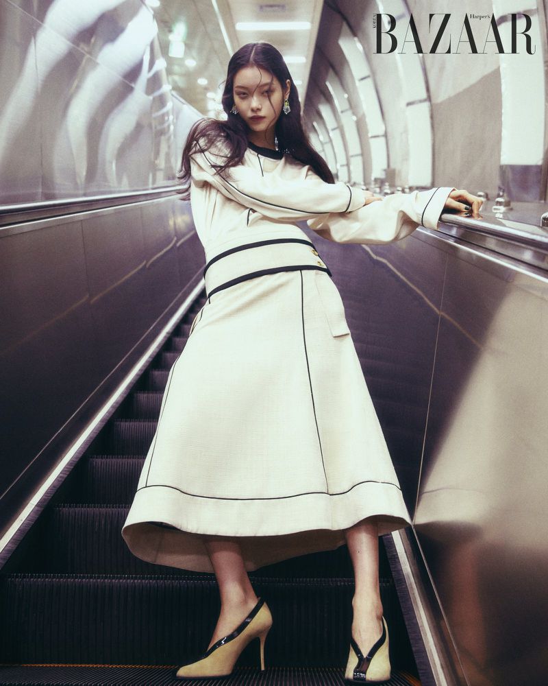 Model standing on a moving escalator wearing an off-white skirt suit with black piping and heels in a subway station.