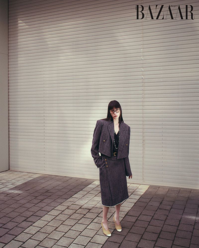 Model standing outdoors wearing a dark tweed suit jacket and skirt set against a slatted metallic wall.
