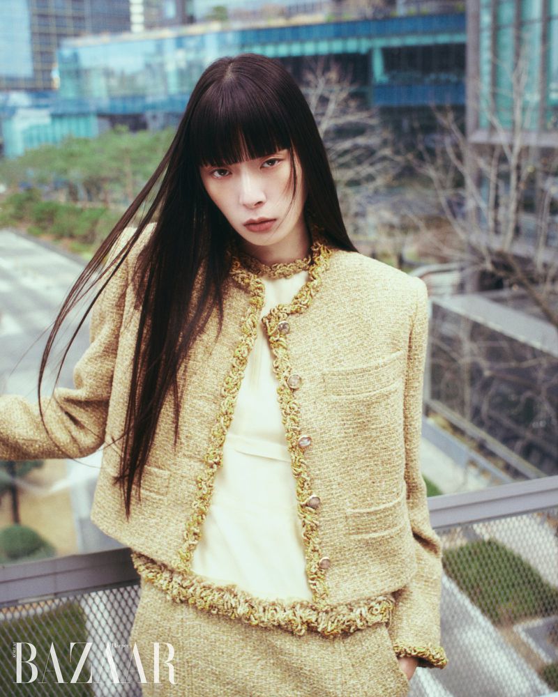 Model with long dark hair wearing a textured tweed jacket standing on a rooftop overlooking buildings