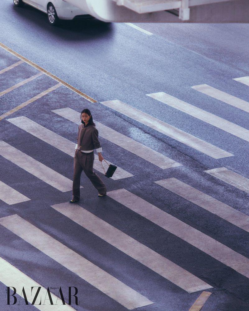 High-angle fashion editorial shot of a model walking across an asphalt crosswalk holding a black handbag.