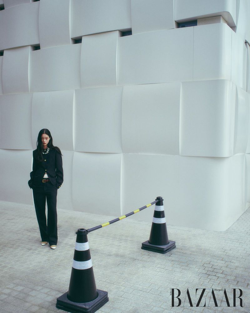Fashion editorial shot of a woman in a black suit standing near black and white striped traffic cones against a white modular wall.
