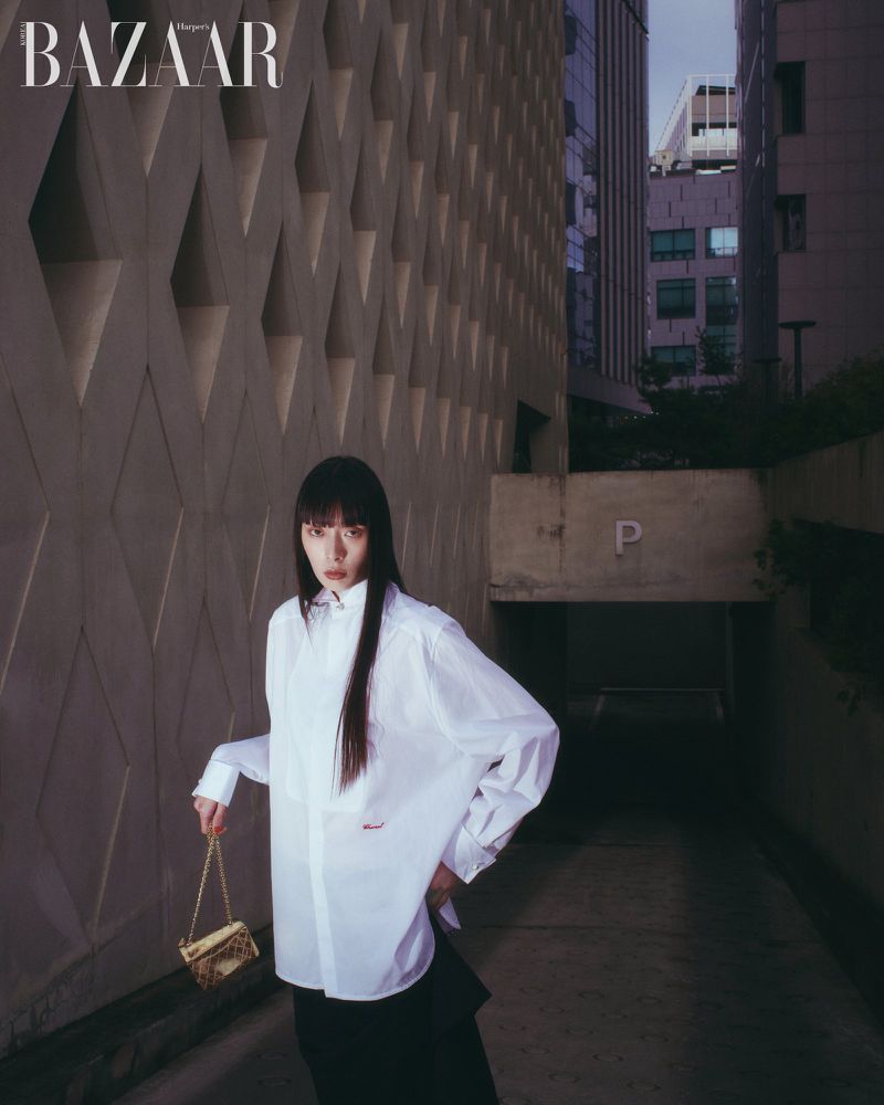 Model with long dark hair wearing a white button-down shirt and holding a gold chain handbag against a patterned concrete wall.