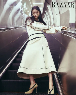 Model standing on a moving escalator wearing an off-white skirt suit with black piping and heels in a subway station.