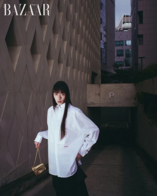 Model with long dark hair wearing a white button-down shirt and holding a gold chain handbag against a patterned concrete wall.