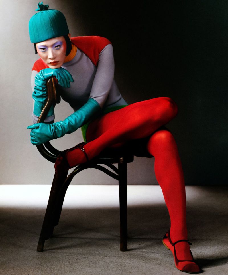 Model Sora Choi wearing a teal beanie, long gloves, and red tights while seated on a dark wooden chair for T Magazine.
