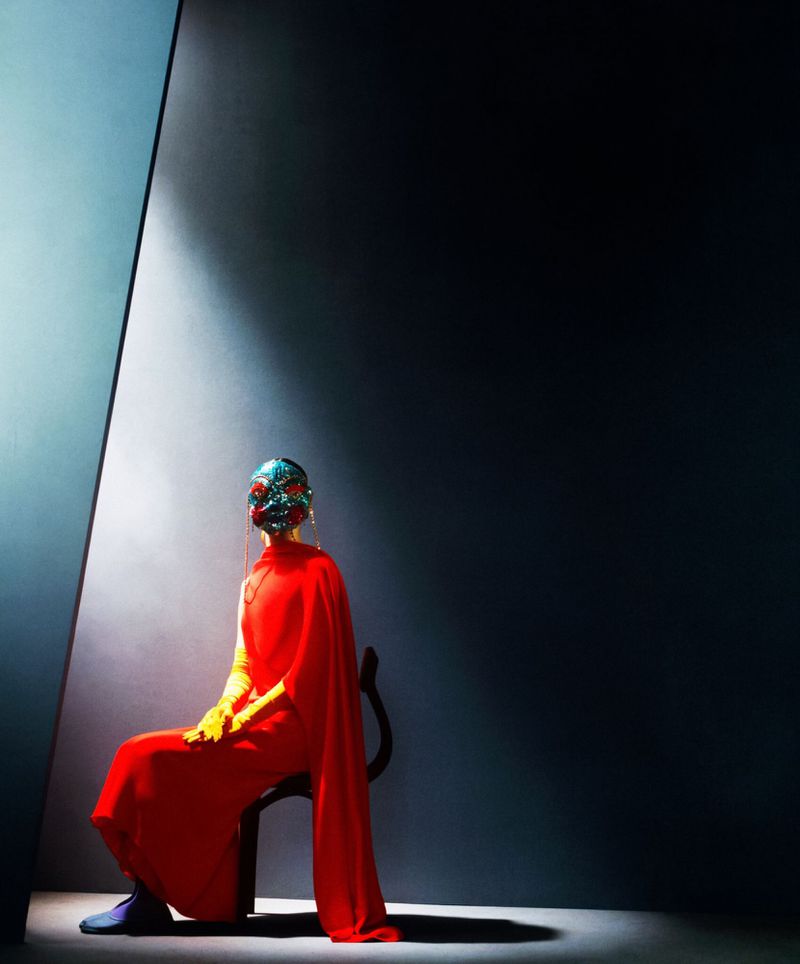 Model wearing a vibrant red dress and ornate mask seated on a dark chair in dramatic moody lighting.
