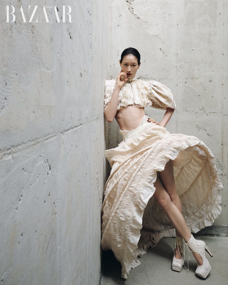 Model wearing a textured cream-colored crop top and ruffled skirt, leaning against a rough concrete wall.