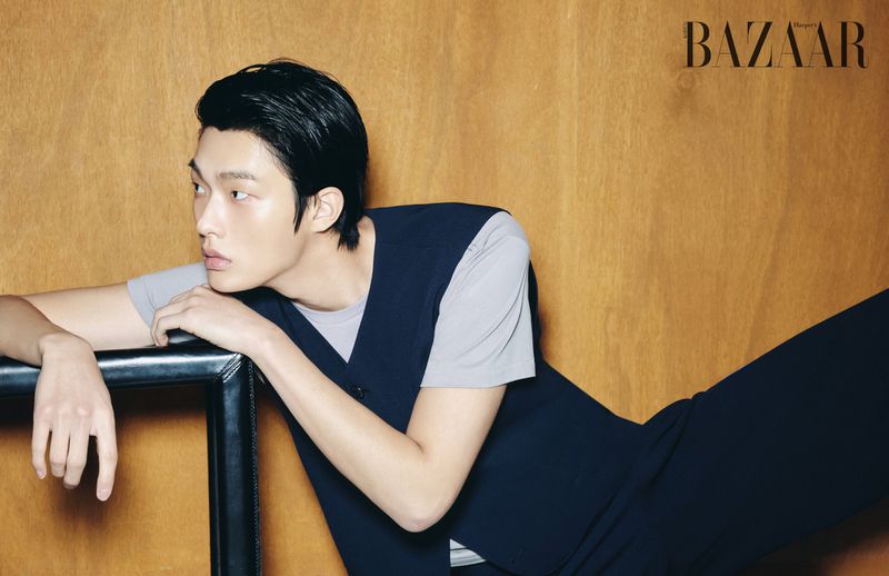 Model Hong Seong Jun wearing a dark vest and grey t-shirt resting against a dark leather frame in front of a warm wood wall.