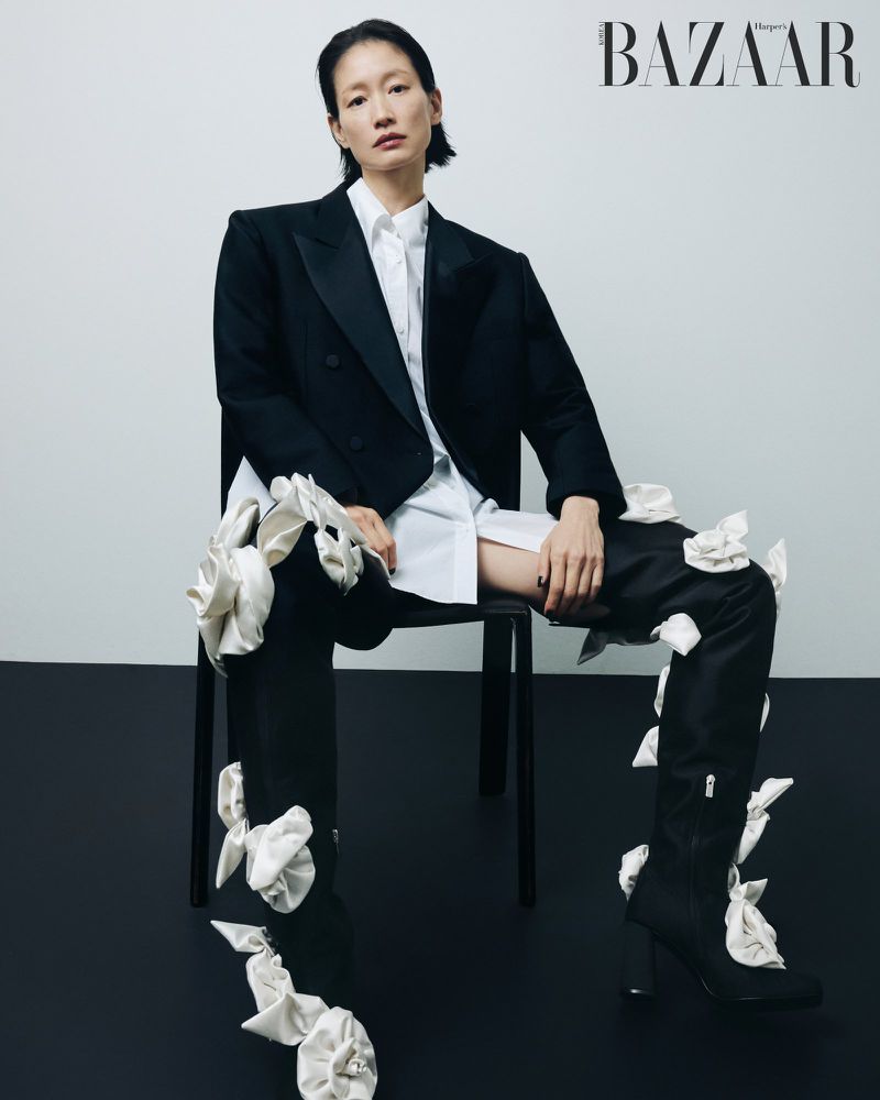Model seated on a black chair wearing a black blazer, white shirt, and embellished tall boots against a light studio backdrop.